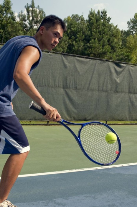 Caught in a mid-backhand swing, this young man was playing a game of tennis on the court. Wearing a darkly-colored tank top, loose-fitting shorts, and a protective layer of sunscreen, all helped block ultraviolet rays from damaging his sun-exposed skin.